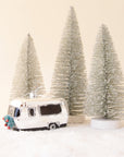 a silver camper ornament with silver bristle trees of varying sizes behind it all sitting on snow with a neutral backgorund