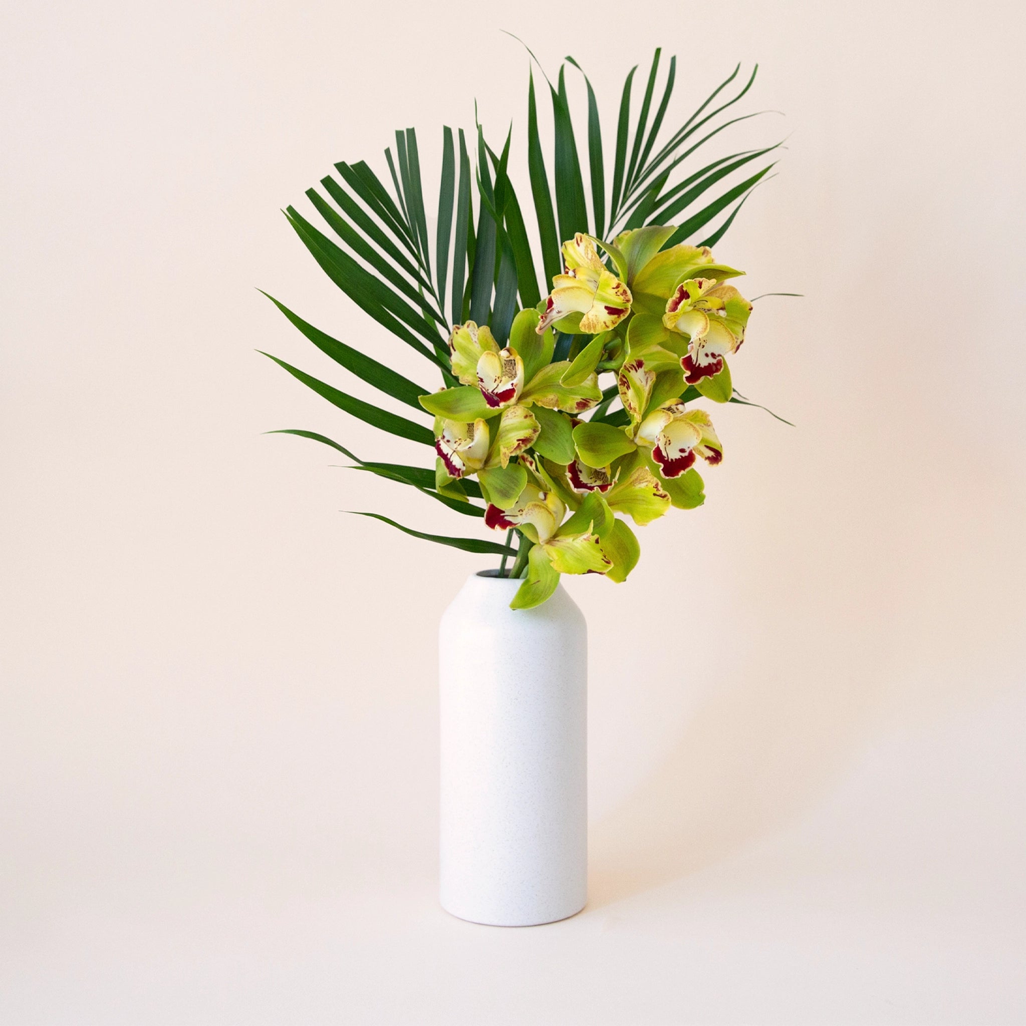 On a cream background is a white ceramic vase with an opening at the top and staged with tropical flowers.