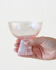 On a white background is a ribbed light pink coupe glass being held by a models hand.