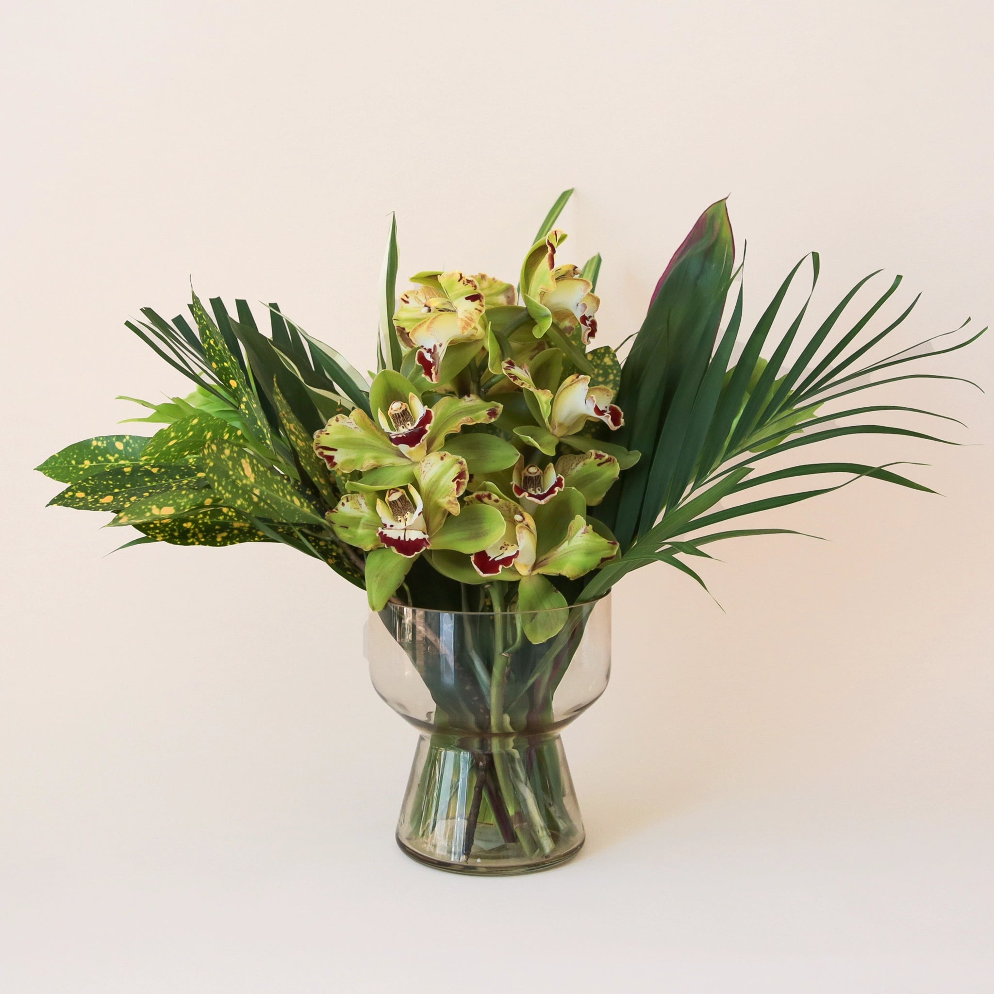On a cream background is a light green glass vase with a green tropical flower arrangement inside that is sold separately. 