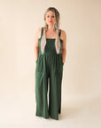 On a cream background is a model wearing a hunter green wide leg jumpsuit with large pockets and 1.5" shoulder straps.