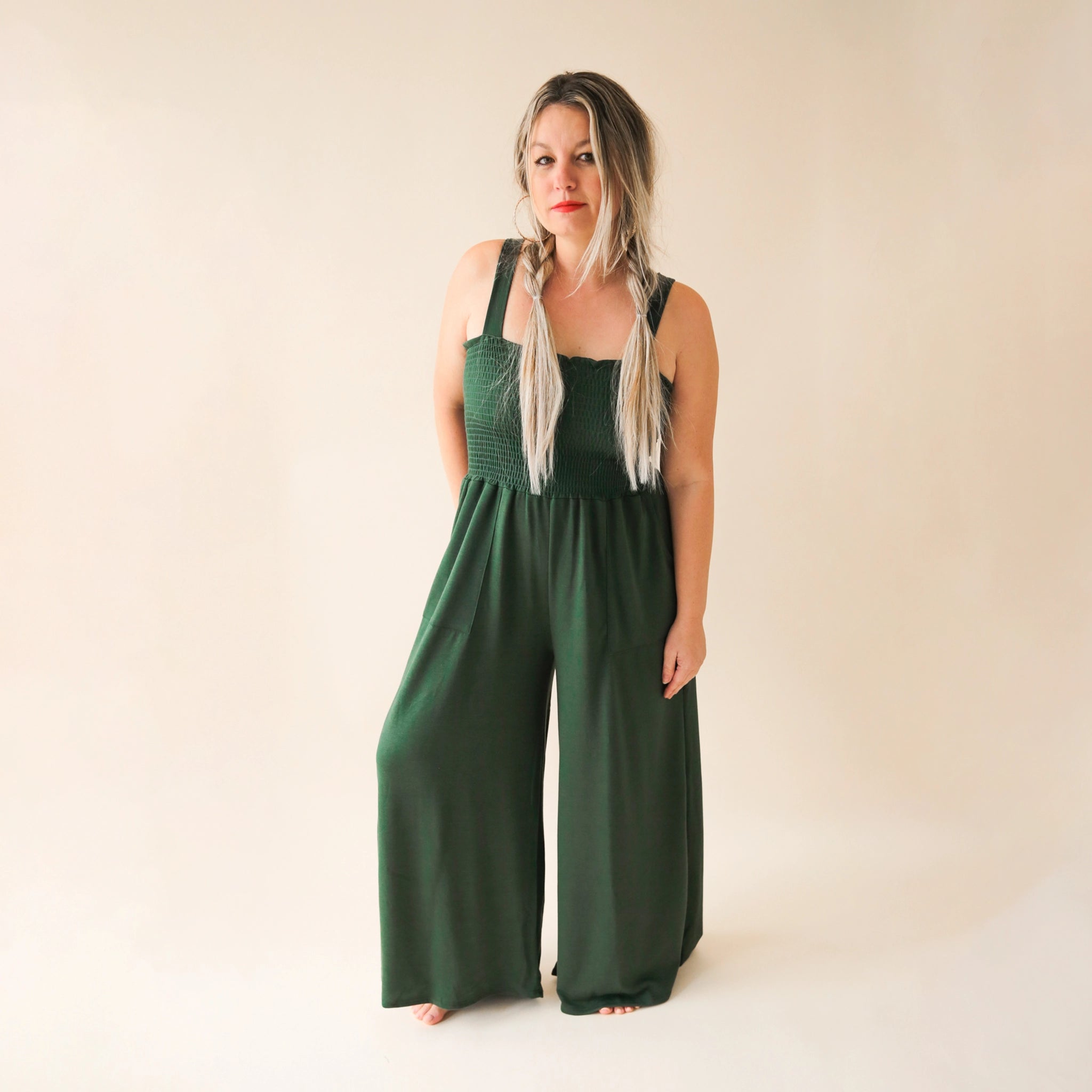 On a cream background is a model wearing a hunter green wide leg jumpsuit with large pockets and 1.5" shoulder straps.