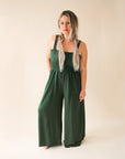 On a cream background is a model wearing a hunter green wide leg jumpsuit with large pockets and 1.5" shoulder straps.