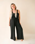 On a cream background is a model wearing a black wide leg jumpsuit with large pockets and 1.5" shoulder straps.