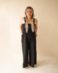 On a cream background is a model wearing a black wide leg jumpsuit with large pockets and 1.5" shoulder straps.