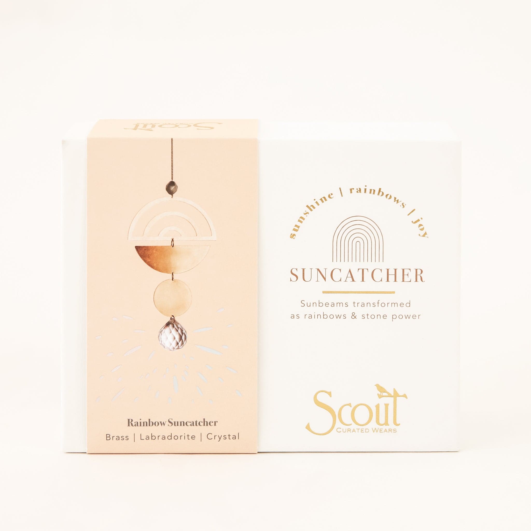 In front of a white background is a white box. On the left of the box is a gold arch of text that reads ‘sunshine | rainbow | joy.’ Under that is more gold text that reads ‘suncatcher.’ At the bottom in gold text it reads ‘Scout.’ On the left side is a peach belly band with a picture of the sun catcher. The clear, round crystal is on the bottom. Above that is a gold, metal circle, then a gold, half circle. Next is another gold, half circle with three arch cut outs. 