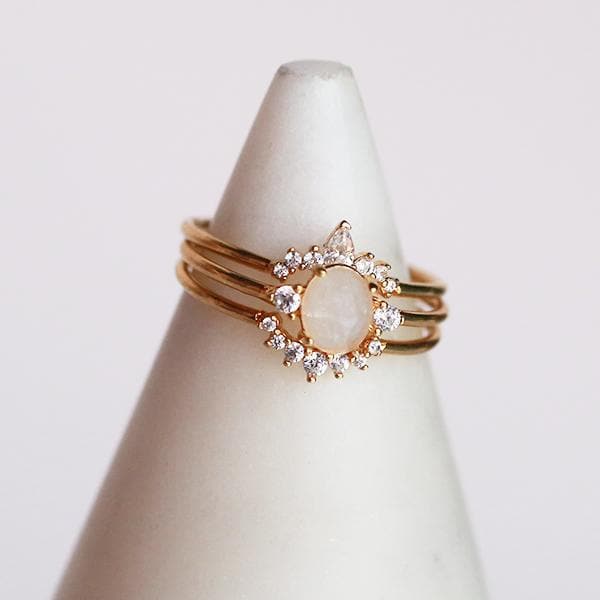In front of a soft pink background and on a white ring holder is three stackable rings. Each ring has a gold band. The top ring has an upward arch in the middle. The arch has three round diamonds on each side and a tall oval diamond in the middle. The middle band has a round white crystal in the middle with a small diamond on each side. The last band has a downward arch with seven small, round diamonds on the arch.