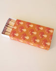 A rectangle box of matches with a repeating red mushroom design along all edges besides the top and bottom. The matches that are inside are also photographed here. They are wooden matches the same length as the box with a red striking tip for lighting.