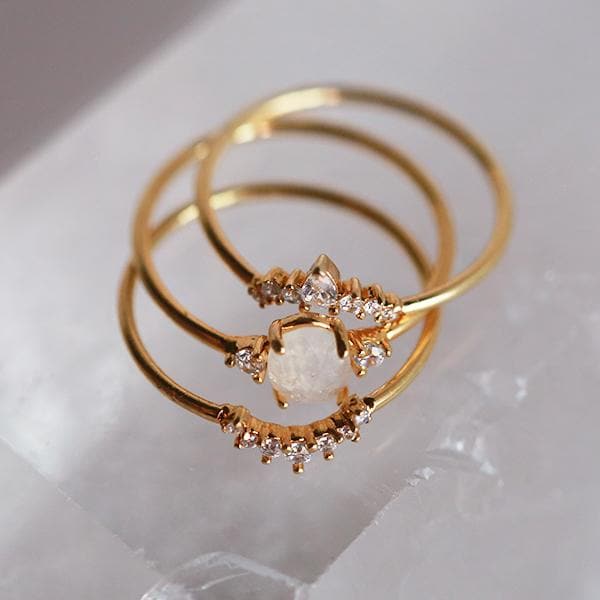 Birds eye view of three stackable rings. Each ring has a gold band. The top ring has an upward arch in the middle. The arch has three round diamonds on each side and a tall oval diamond in the middle. The middle band has a round white crystal in the middle with a small diamond on each side. The last band has a downward arch with seven small, round diamonds on the arch.