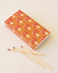 A rectangle box of matches with a repeating red mushroom design along all edges besides the top and bottom. The matches that are inside are also photographed here. They are wooden matches the same length as the box with a red striking tip for lighting.