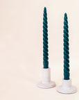Against a white background are two gray, ceramic candle holders. The candle holder on the left has a round base and tall, cylinder holder. The holder on the left also has a round base but the cylinder holder is about half the size of the other holder. Both holders have a dark green, twisted taper candle inside.