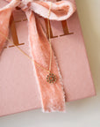 A dainty gold chain necklace with a gold circle pendant in the center with a cut out of a daisy flower, photographed on a pink book with a textured pink ribbon.