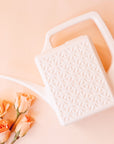 On a peachy background is a white plastic watering can with a long curved spout and a squared off handle along with a breeze block textured design on the sides of the watering can photographed next to peachy colored roses.