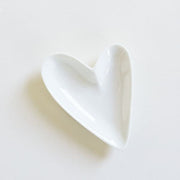 On a white background is a white heart shaped ring dish with a glazed ceramic finish.