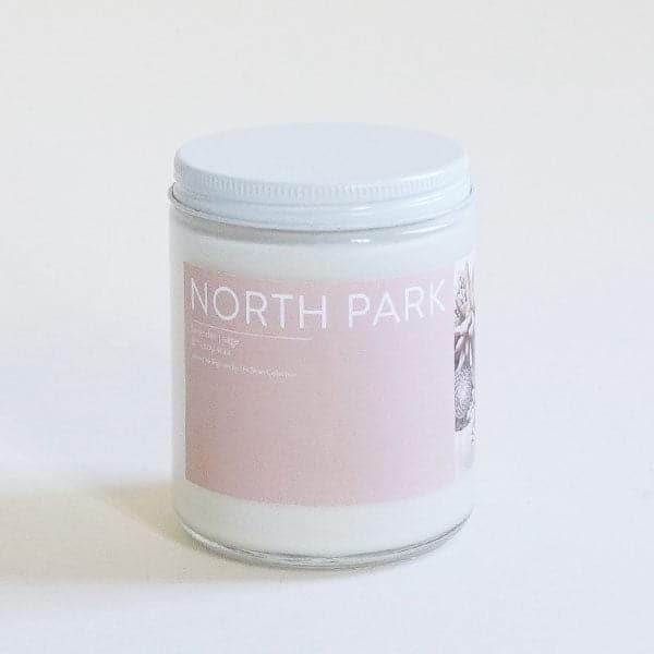 On a white background is a clear glass jar candle with a light pink label on the front that reads, "North Park" along with a white screw on lid. 
