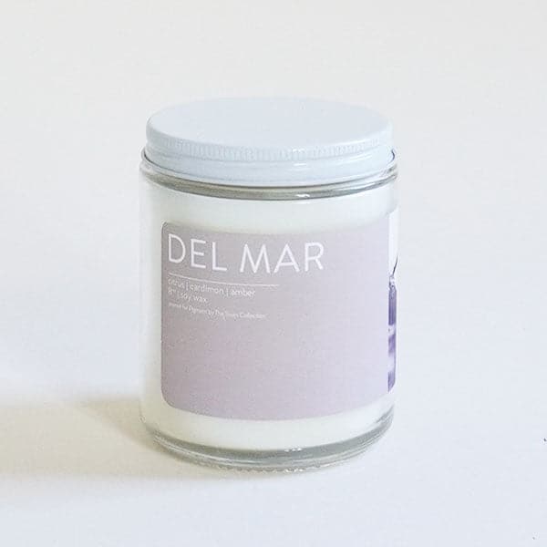 A clear glass jar candle with a light lavender label with white text that reads, "Del Mar" and a white lid.