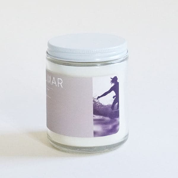 A clear glass jar candle with a light lavender label with white text that reads, "Del Mar" and a white lid and a rectangular photo of a surfer.
