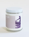 A clear glass jar candle with a light lavender label with white text that reads, "Del Mar" and a white lid and a rectangular photo of a surfer.