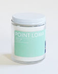 In front of a white background is a clear glass cylinder jar. The jar has a white top. On the front of the jar is a turquoise sticker. On the top of the sticker is white text that reads ‘Point Loma.'