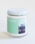 In front of a white background is a clear glass cylinder jar. The jar has a white top. On the front of the jar is a turquoise sticker. On the right side is a small picture of a VW bus in front of a blue sky.