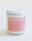 In front of a white background is a clear glass, round jar with a white metal lid. On the front is a pink sticker with white text at the top that reads ‘San Diego.'
