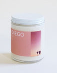 In front of a white background is a clear glass, round jar with a white metal lid. On the front is a pink sticker with white text at the top that reads ‘San Diego.’ On the left side of the sticker is a small picture of a dark pink and yellow sunset with a couple palm trees.