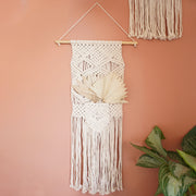 On a terracotta colored wall is a natural colored macrame wall hanger wrapped on a wood dowel and detailed with tassels along the bottom edge and a pocket in the front.