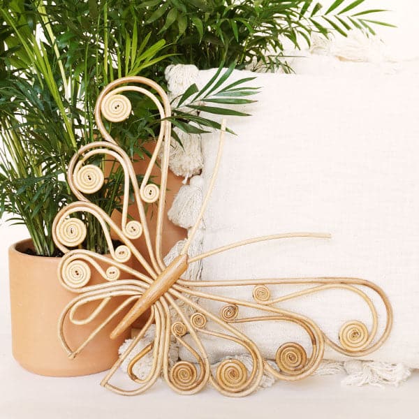 Rattan shaped butterfly wall art with curled wings staged in front of plants and a cream colored pillow.