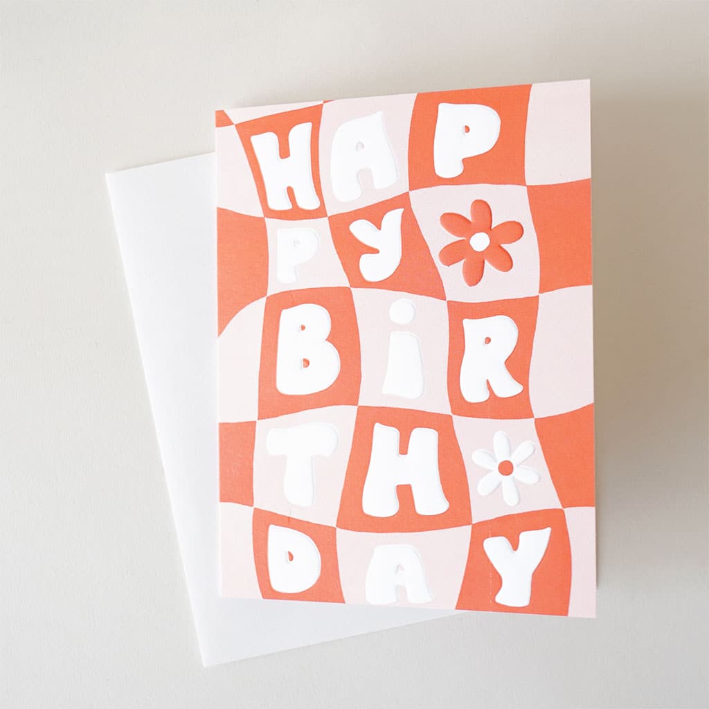 Card reading 'Happy Birthday' against a trippy pastel pink and deep orange checkerboard. Two daisies are featured within the checkered pattern on the right hand side. The card is accompanied by a solid white envelope. 