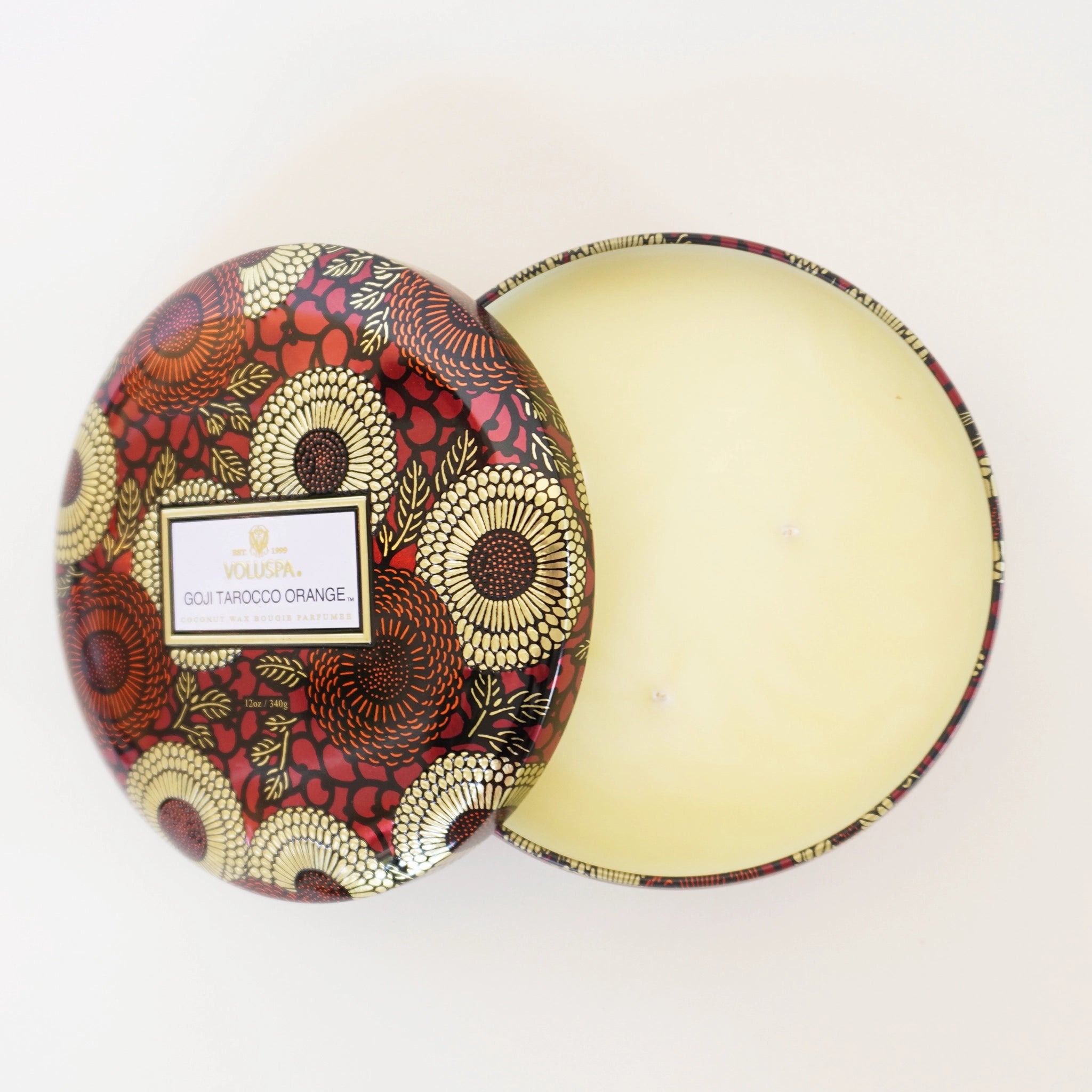 On a white background is a red tin with a three wick candle inside and a label on the lid that reads, &quot;Voluspa Goji Torocco Orange&quot;.