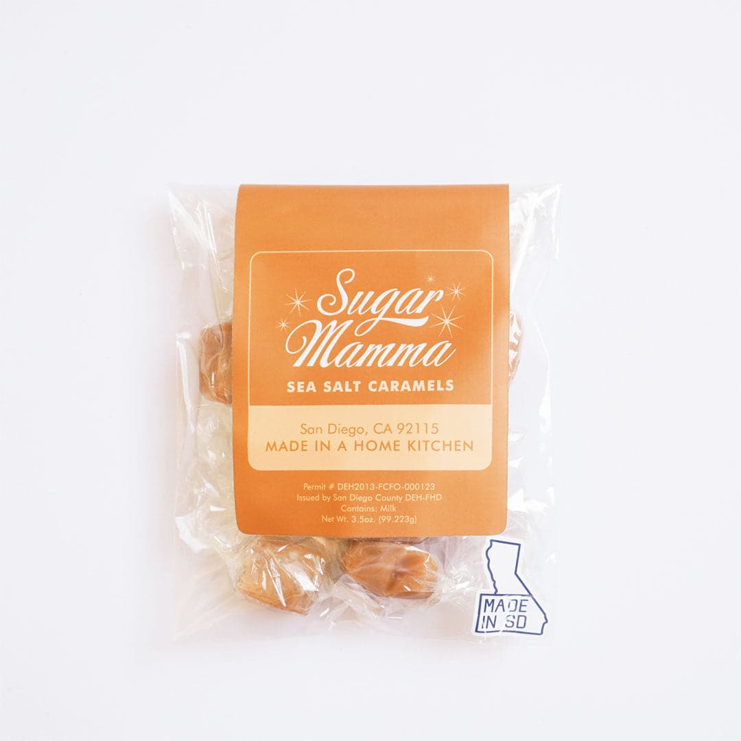 This is a clear, square plastic package. Inside the package is light brown candies. There is a light orange vertical belly band that stops before touching the bottom. In the middle of the belly band is a white outlined square. Inside is white script that reads ‘sugar mamma.’ Below is more white text that reads ‘sea salt caramels.’ The bottom half of the square is peach with light orange text that reads ‘San Diego, CA 92115 Made in a home kitchen.’ 