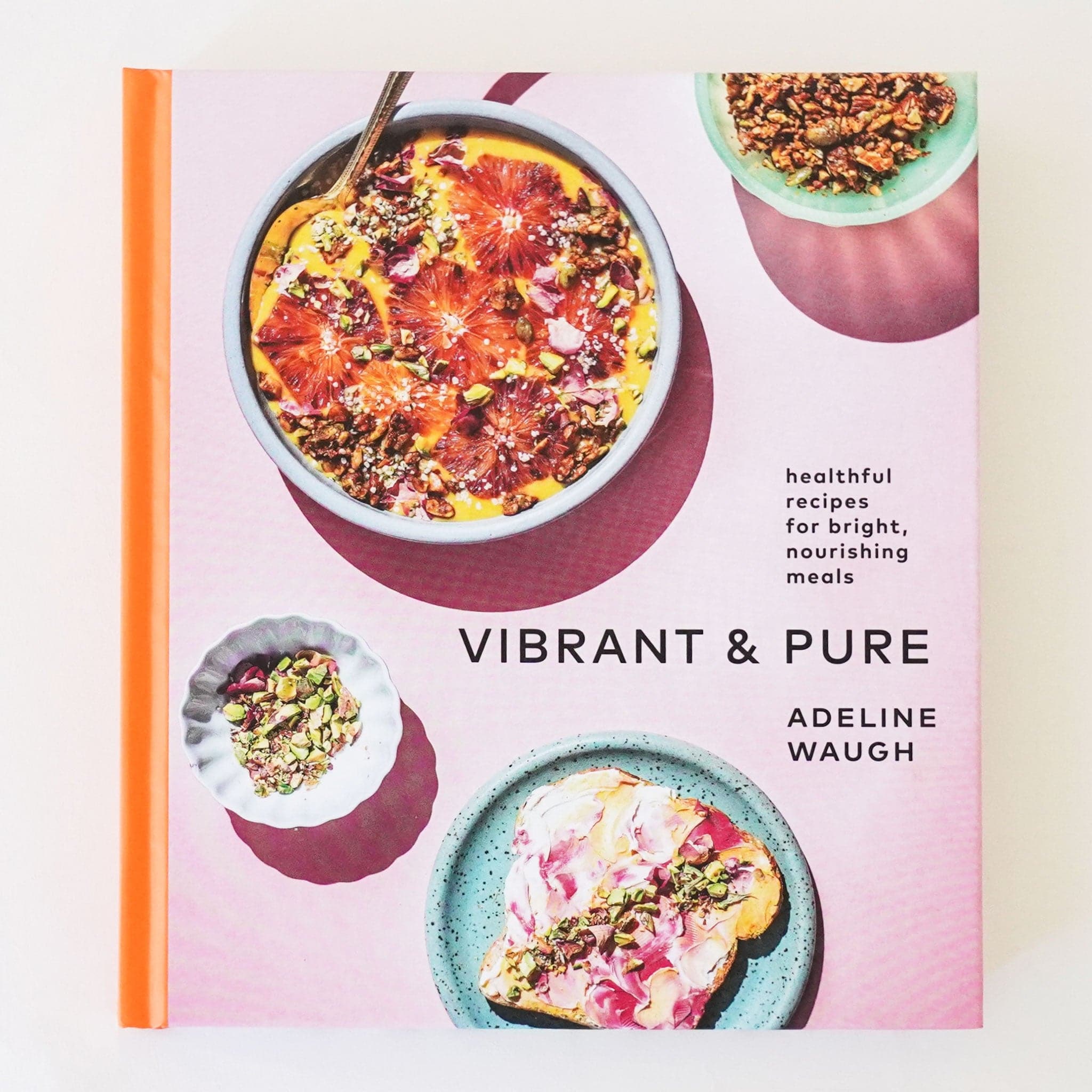 Front cover of cookbook reading 'healthful recipes for bright, nourishing meals, vibrant & Pure'. The cover is a soft lilac with tangerine orange binding. Pictured around the text are smoothie bowl, plated toast and more. 