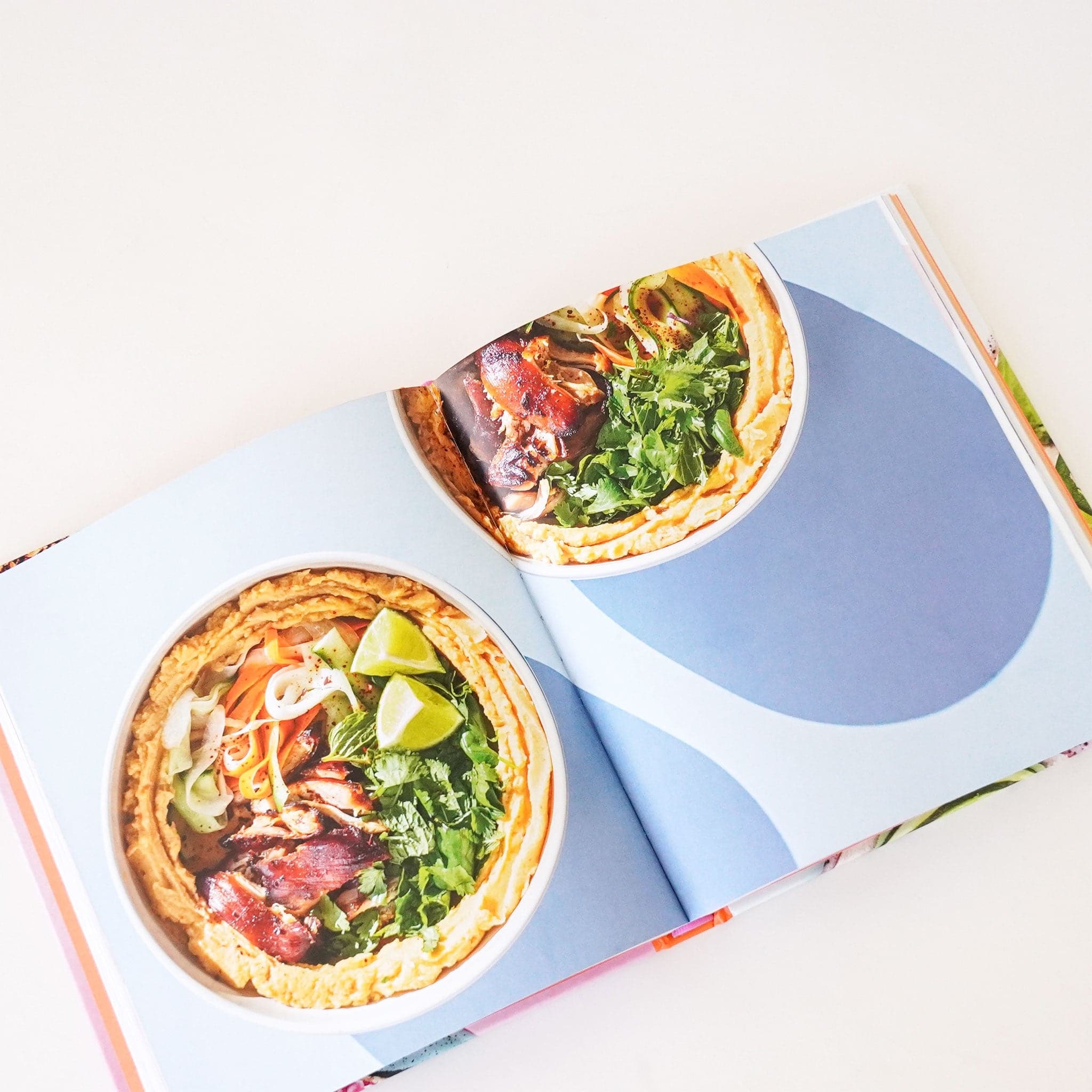 Open pages of book featuring two vibrantly colorful bowls of salads against a baby blue background. 