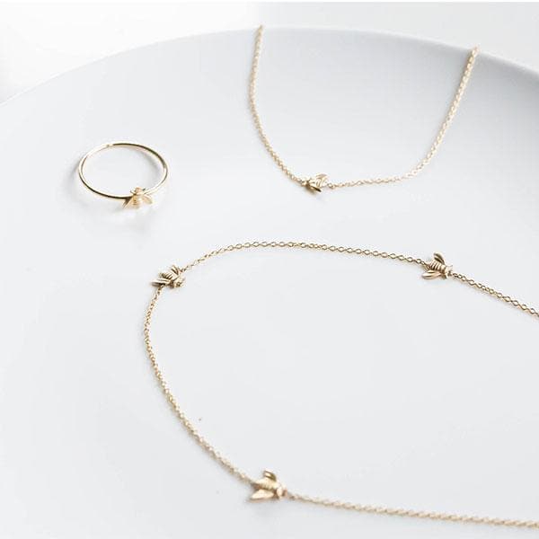 A dainty thin gold band ring with a small gold bee in the center, photographed next to a coordinating bee necklaces.