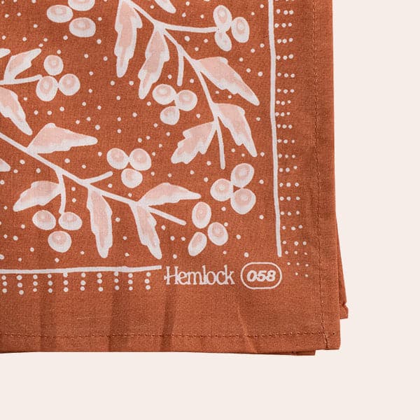 Against a white background is a dark terracotta bandana. This is a close up of the pink and white leaves and flowers pattern. There are white polka dots surrounding the flowers. In the bottom corner there is white text that reads ‘hemlock.’ 