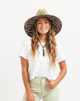 In front of a white background is a woman wearing a white t-shirt and tan pants. On her head she is wearing a wide brim, straw hat. The brim of the hat has a black and brown cheetah print border. It also has a black drawstring.