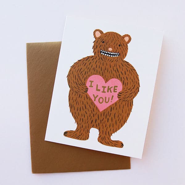 A white folded card with a brown bear graphic on the front holding a pink heart that says, &quot;I Like You&quot; in black writing.