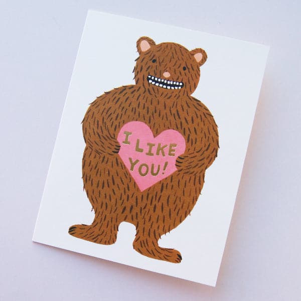 A white folded card with a brown bear graphic on the front holding a pink heart that says, "I Like You" in black writing.