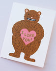 A white folded card with a brown bear graphic on the front holding a pink heart that says, "I Like You" in black writing.