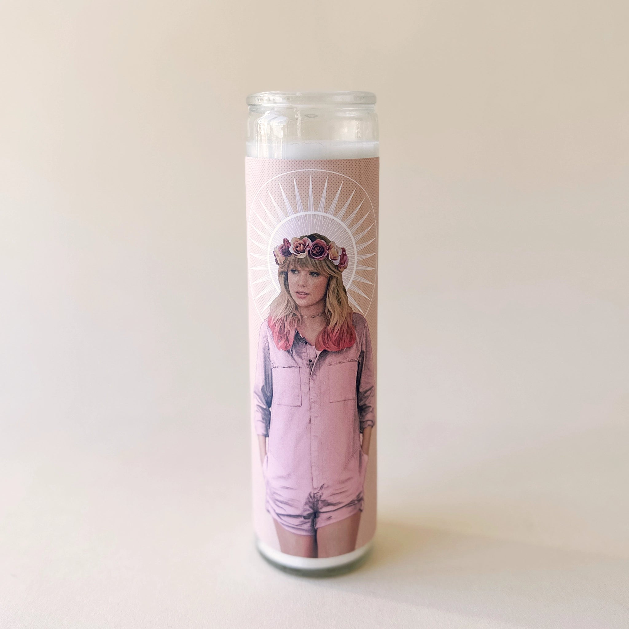 A thin prayer candle in a glass jar with a pink image of Taylor Swift in a pink jumpsuit with a rose flower crown on.