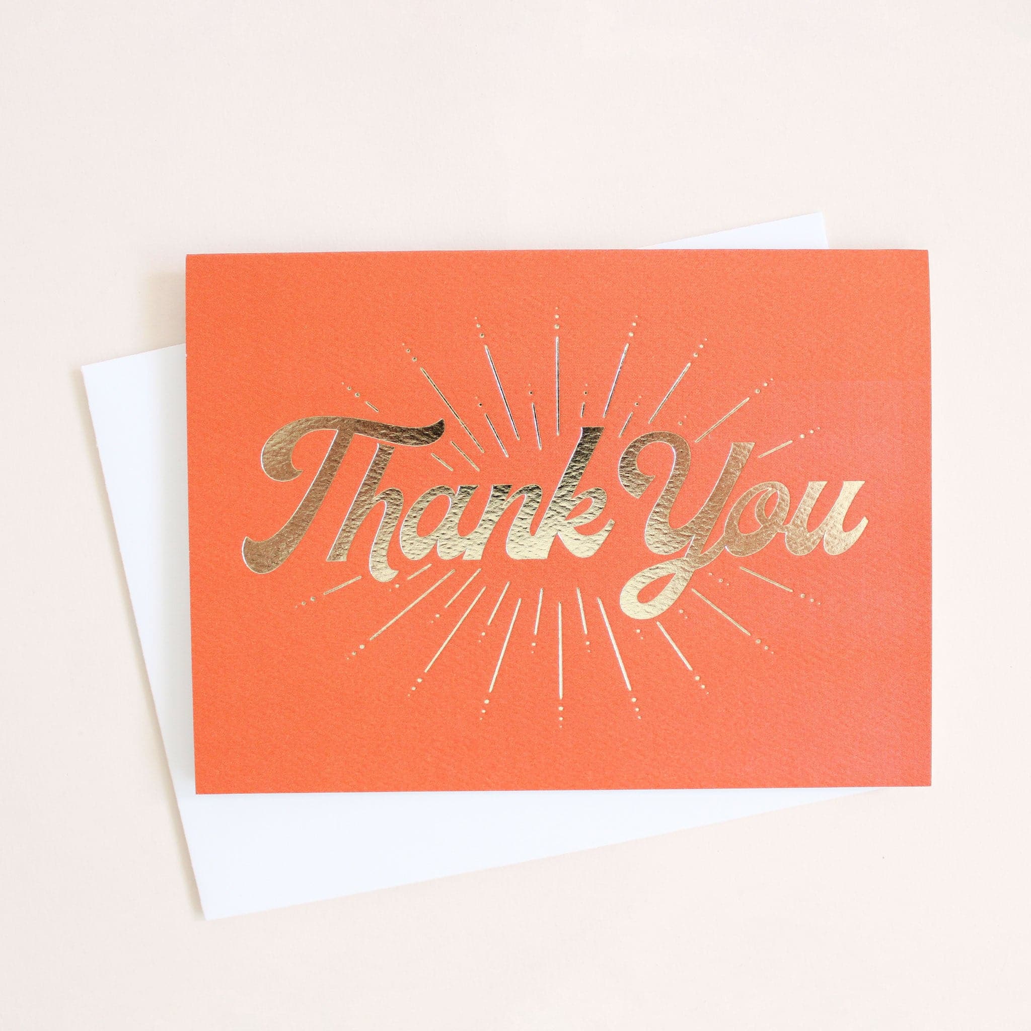 Tangerine orange card reading &#39;Thank You&#39; in gold foil. Gold foil sunburst design beams from the text. The card is accompanied by a solid white envelope