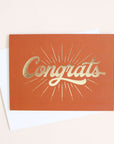 deep orange colored greeting card with a gold foil starburst design surrounding the word congrats.
