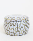 Against a white background is the side view of a round tin candle with a matching lid. The candle and the lid are white with a gold and silver design.