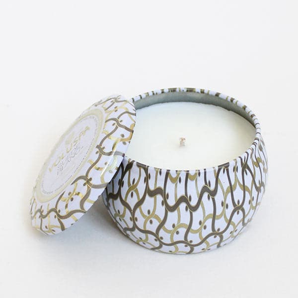 Against a white background is the side view of a round tin candle with a matching lid leaning against the left side of the candle. The candle and the lid are white with a gold and silver design. Inside the tin is a white candle. 