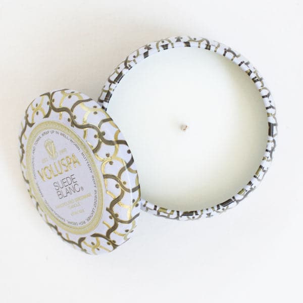 Against a white background is the birds eye view of a round, tin candle. The border of the candle is white with silver and gold lines. The candle inside the tin is white with a white wick. A matching tin lid is laying against the left side of the candle. In the middle is a gold circle with white inside. In gold text it reads ‘Voluspa.’ Below is black text that reads ‘suede blanc.’ 