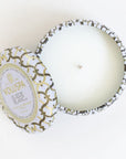 Against a white background is the birds eye view of a round, tin candle. The border of the candle is white with silver and gold lines. The candle inside the tin is white with a white wick. A matching tin lid is laying against the left side of the candle. In the middle is a gold circle with white inside. In gold text it reads ‘Voluspa.’ Below is black text that reads ‘suede blanc.’