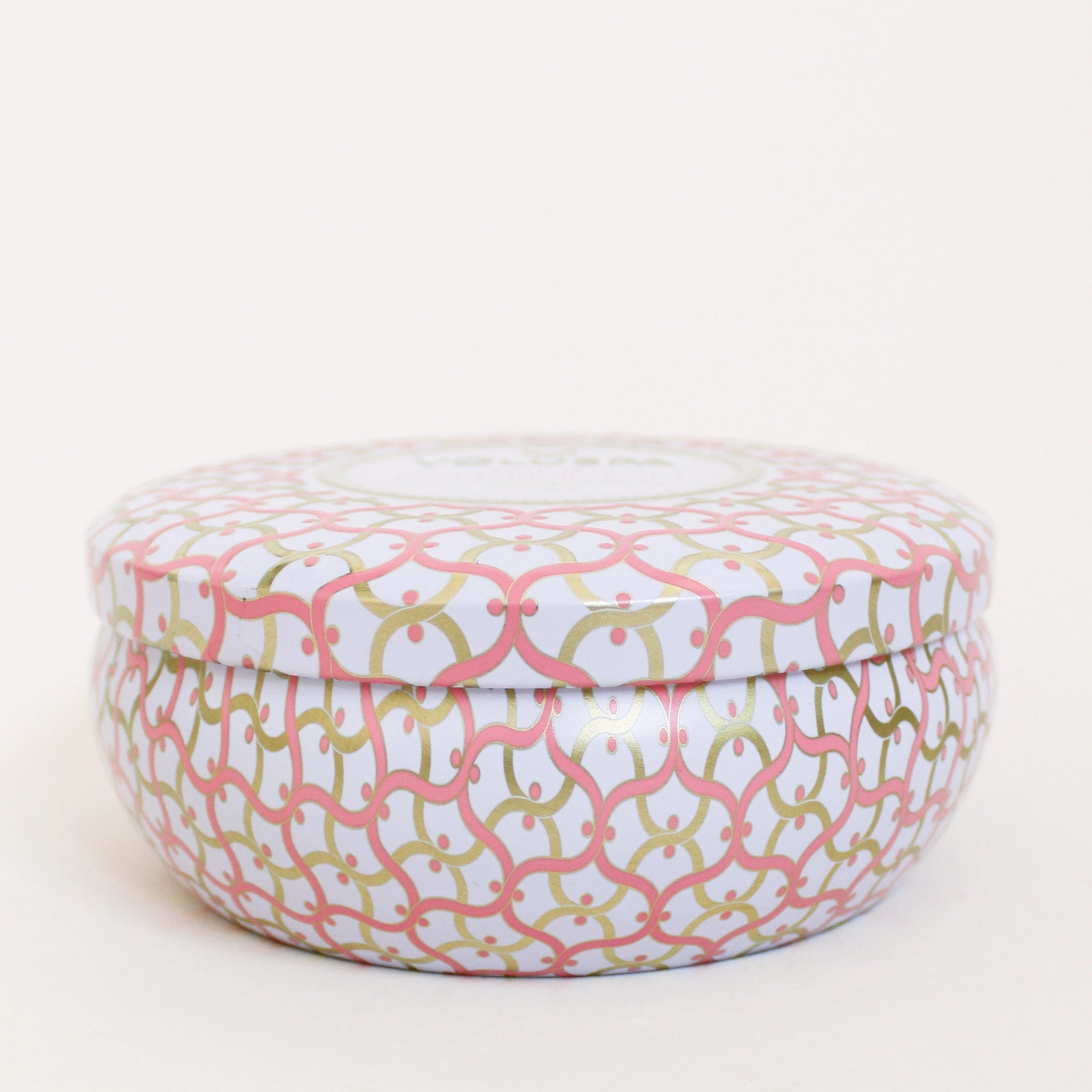 In front of a white background is a round tin candle. The tin is white with pink and gold swirly lines. There is a matching lid on top.