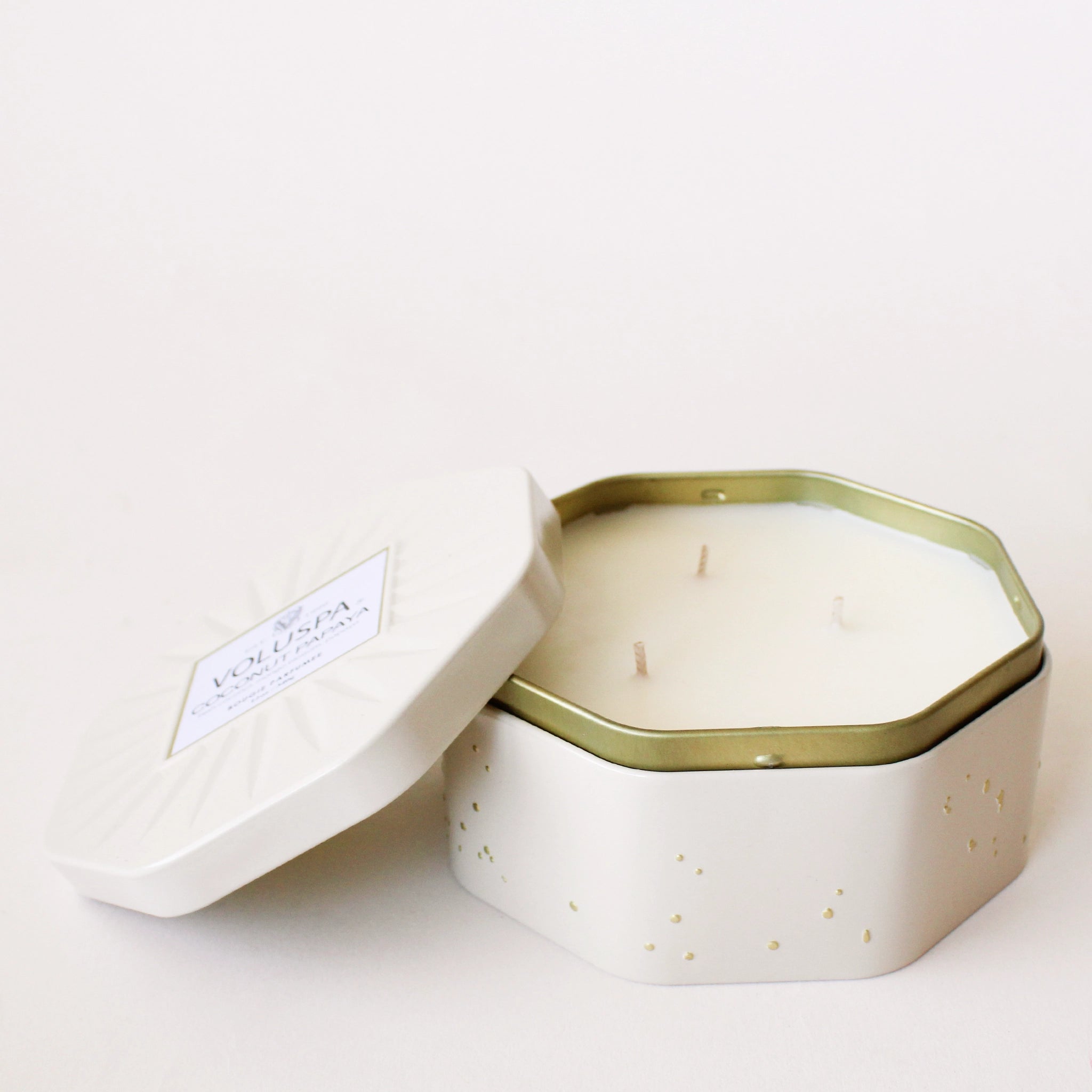 On a white background is an octagon tin in a white shade with a white three wick candle inside along with a label on the lid that reads, &quot;Voluspa Coconut Papaya&quot;.