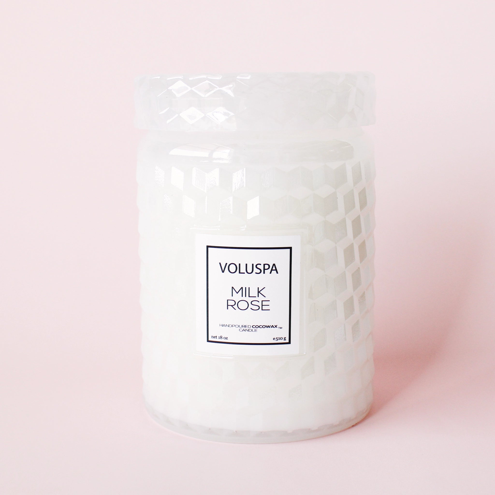 On a light pink background is a white glass jar candle with a milky effect to the glass along with a lid and a rectangular label in the center that reads, "Voluspa Milk Rose".