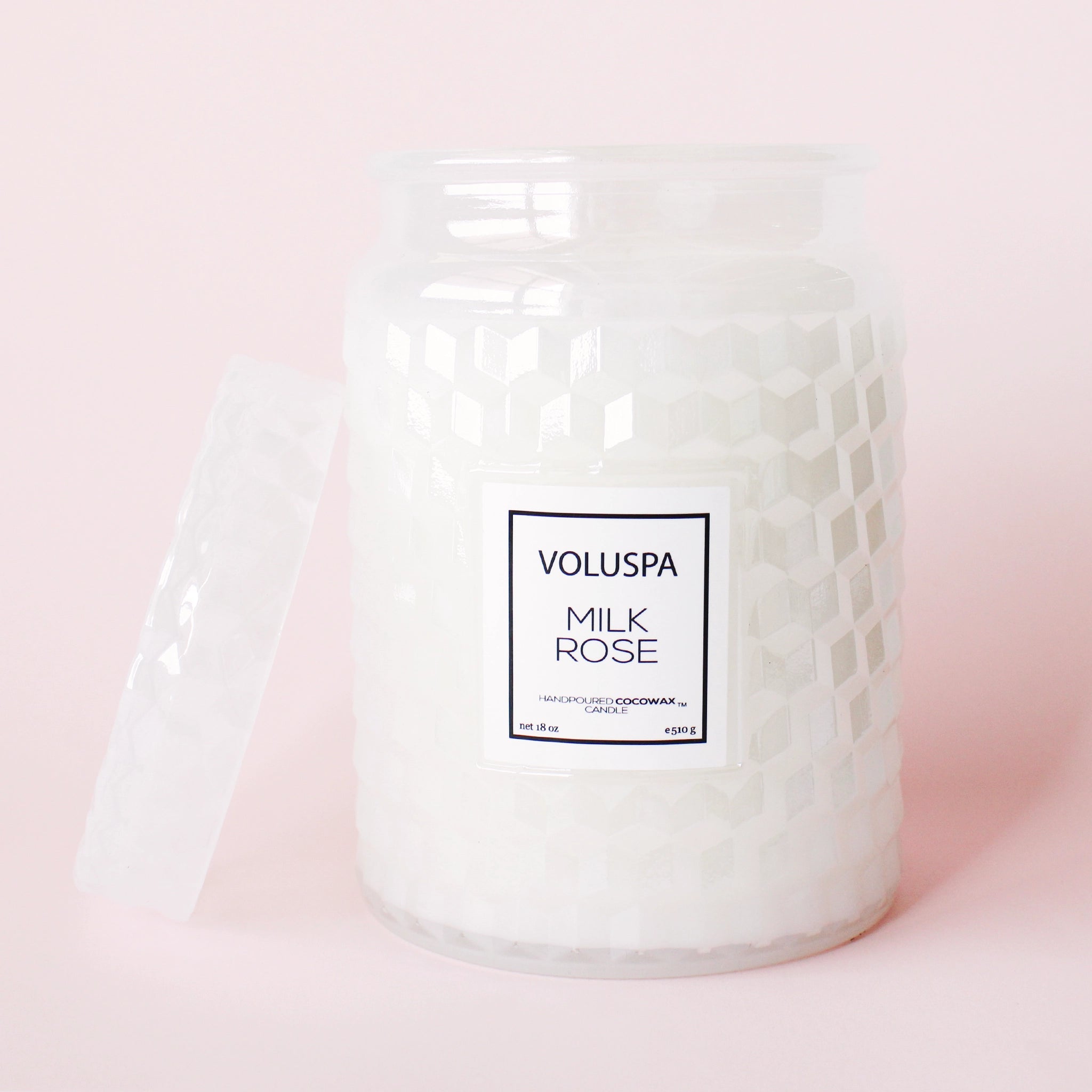 On a light pink background is a white glass jar candle with a milky effect to the glass along with a lid and a rectangular label in the center that reads, &quot;Voluspa Milk Rose&quot;.